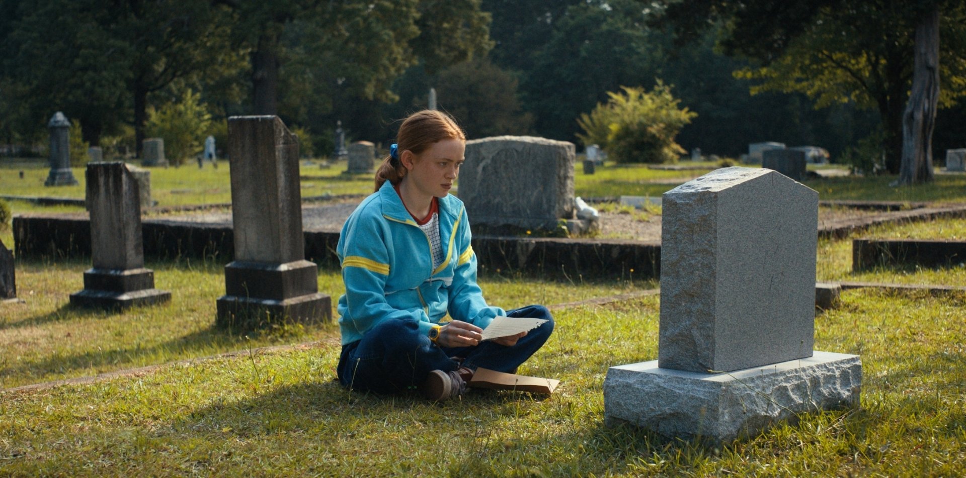 Young woman in a blue-and-yellow jacket sits cross-legged in a cemetery reading beside a gravestone — HD PC desktop wallpaper evoking a TV drama character.