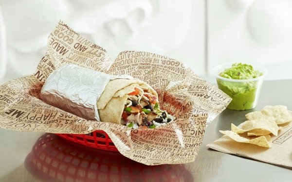 Freshly made burrito wrapped in branded paper beside a bowl of guacamole and tortilla chips, set against a blurred background, perfect for HD desktop wallpaper or background.