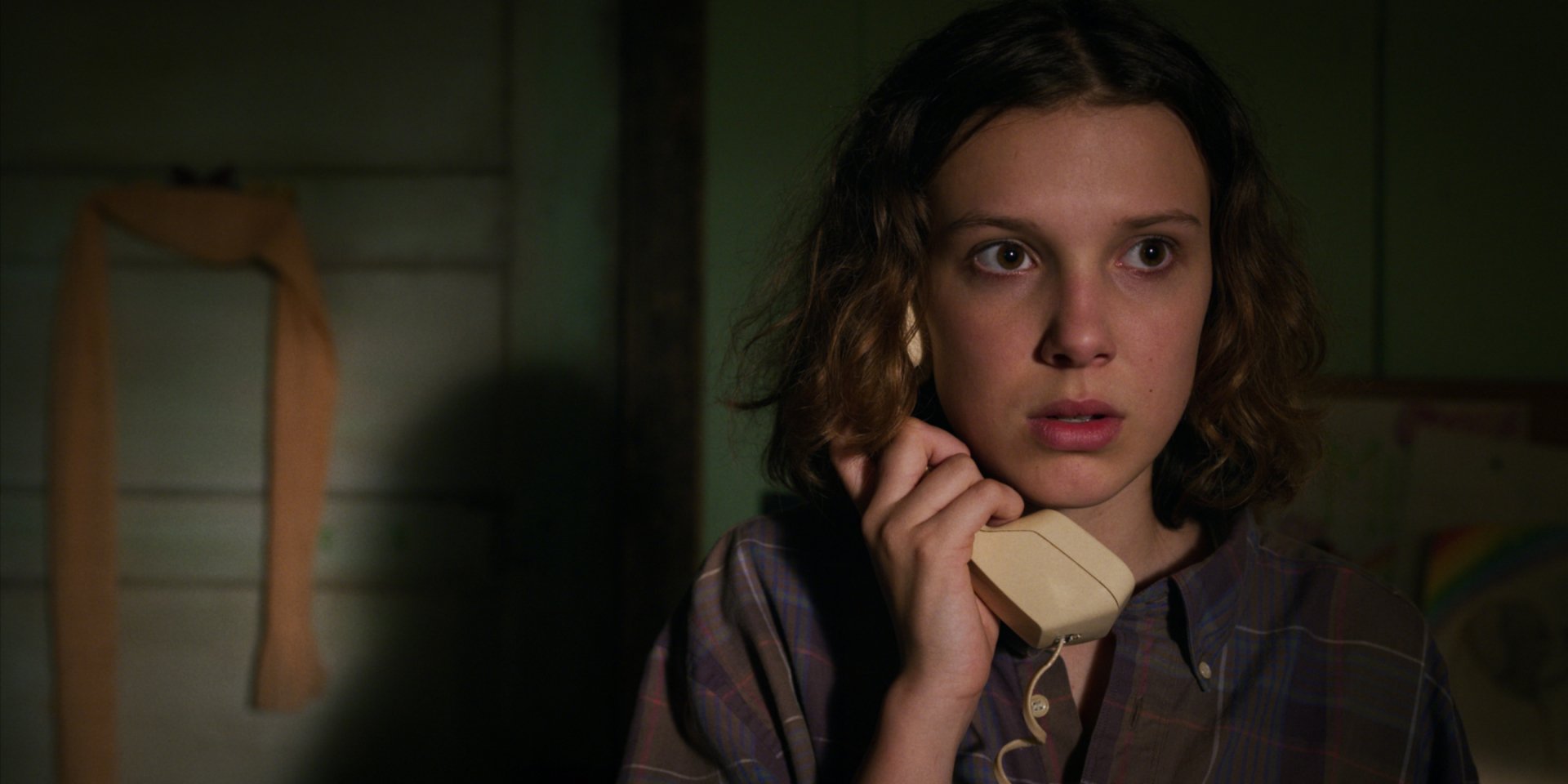 Eleven from Stranger Things, portrayed by Millie Bobby Brown, shown in a tense moment holding a phone in this HD TV show desktop wallpaper and background.