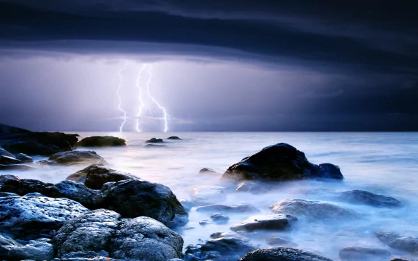 HD desktop wallpaper showing a stormy ocean scene with dark clouds, lightning striking over the water, and rocky shore in the foreground, highlighting dramatic natural power.