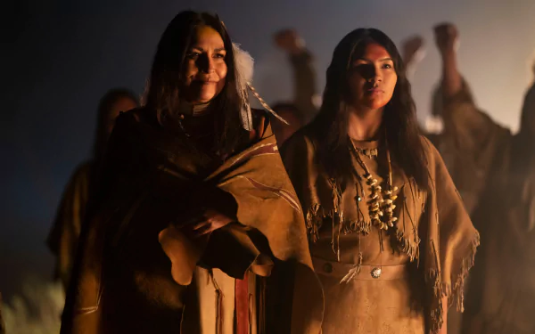 Two women in traditional leather garments stand in warm firelight during a tense scene from the movie Prey (2022). 2K Quad HD PC desktop wallpaper/background.