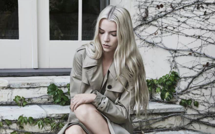 HD PC desktop wallpaper: blonde female celebrity seated on stone steps wearing a trench coat, contemplative expression, with ivy-covered wall and window behind her.