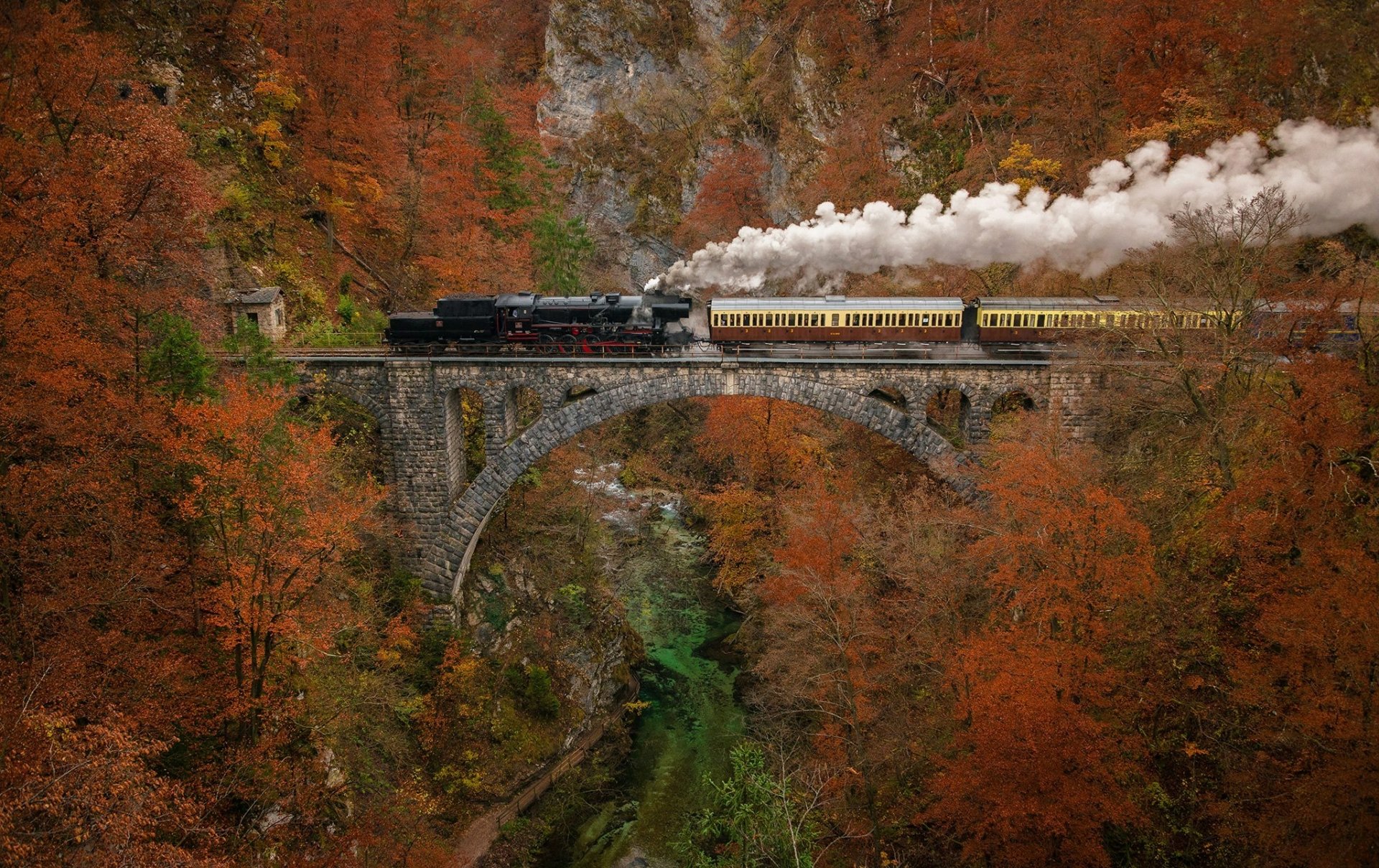 A vintage train travels across a stone bridge surrounded by vibrant fall foliage, creating a stunning HD desktop wallpaper scene.