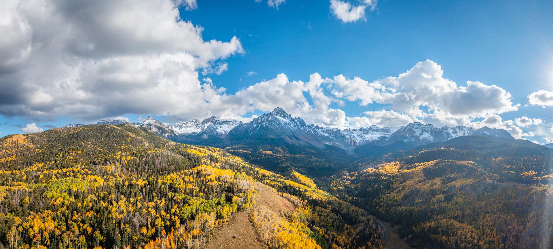 A stunning 4K Ultra HD nature landscape showcasing snow-capped mountains under a vibrant blue sky with scattered clouds and golden autumn trees.