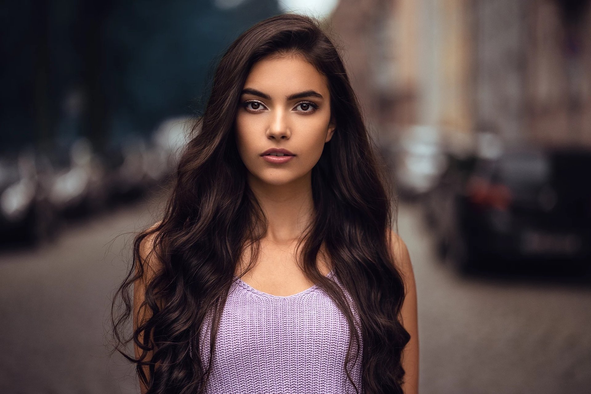 HD desktop wallpaper featuring a confident woman model with long dark hair, wearing a lavender top, standing outdoors on a blurred street background.