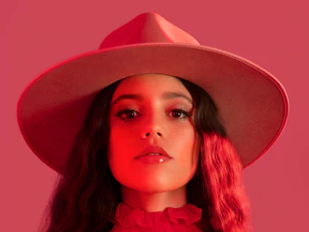 HD PC desktop wallpaper featuring celebrity Jenna Ortega in a wide-brimmed hat against a red background.