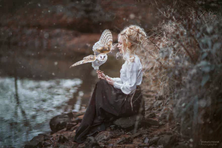 HD PC desktop wallpaper of a woman with flowing hair sitting by water, gently holding a barn owl, evoking a serene and introspective mood.