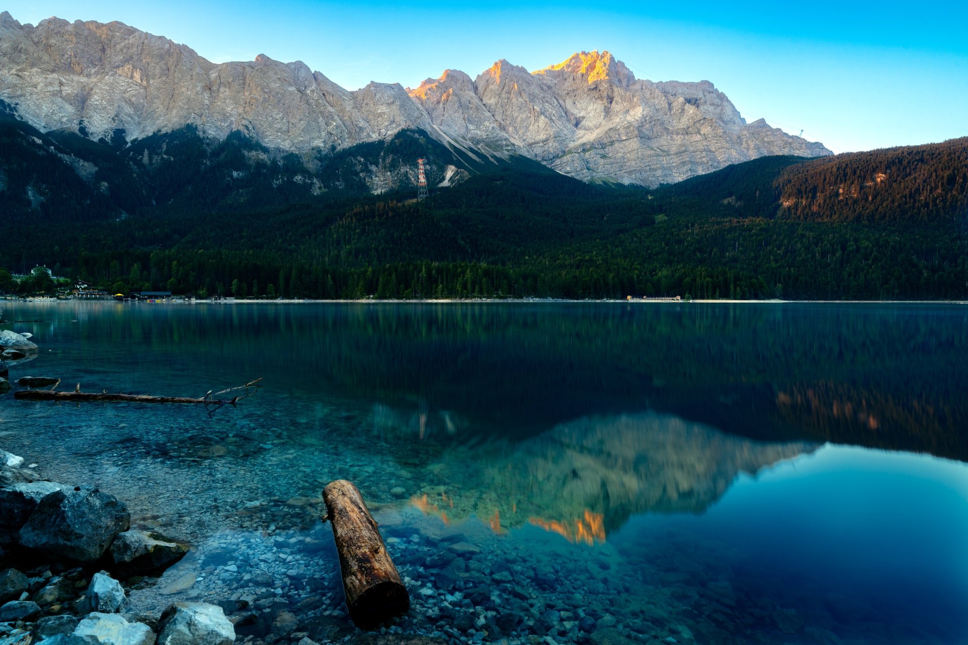 An 8K Ultra HD nature wallpaper featuring a serene mountain lake with crystal-clear water reflecting the towering peaks and a tranquil sky.