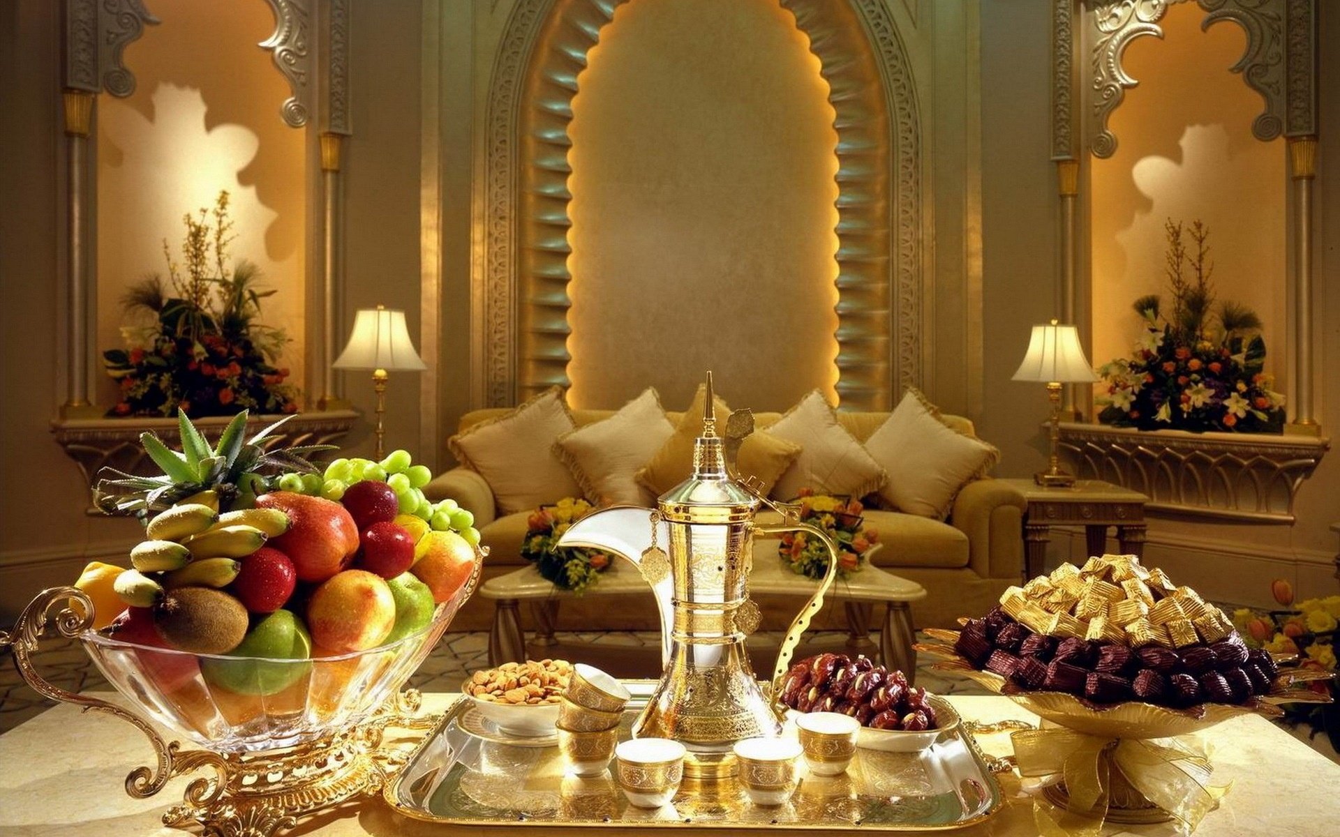 HD PC desktop wallpaper background: opulent still-life food scene in a Middle Eastern lounge — brass coffee pot on a tray, bowls of fruit, dates and sweets on a polished table before an ornate arch.