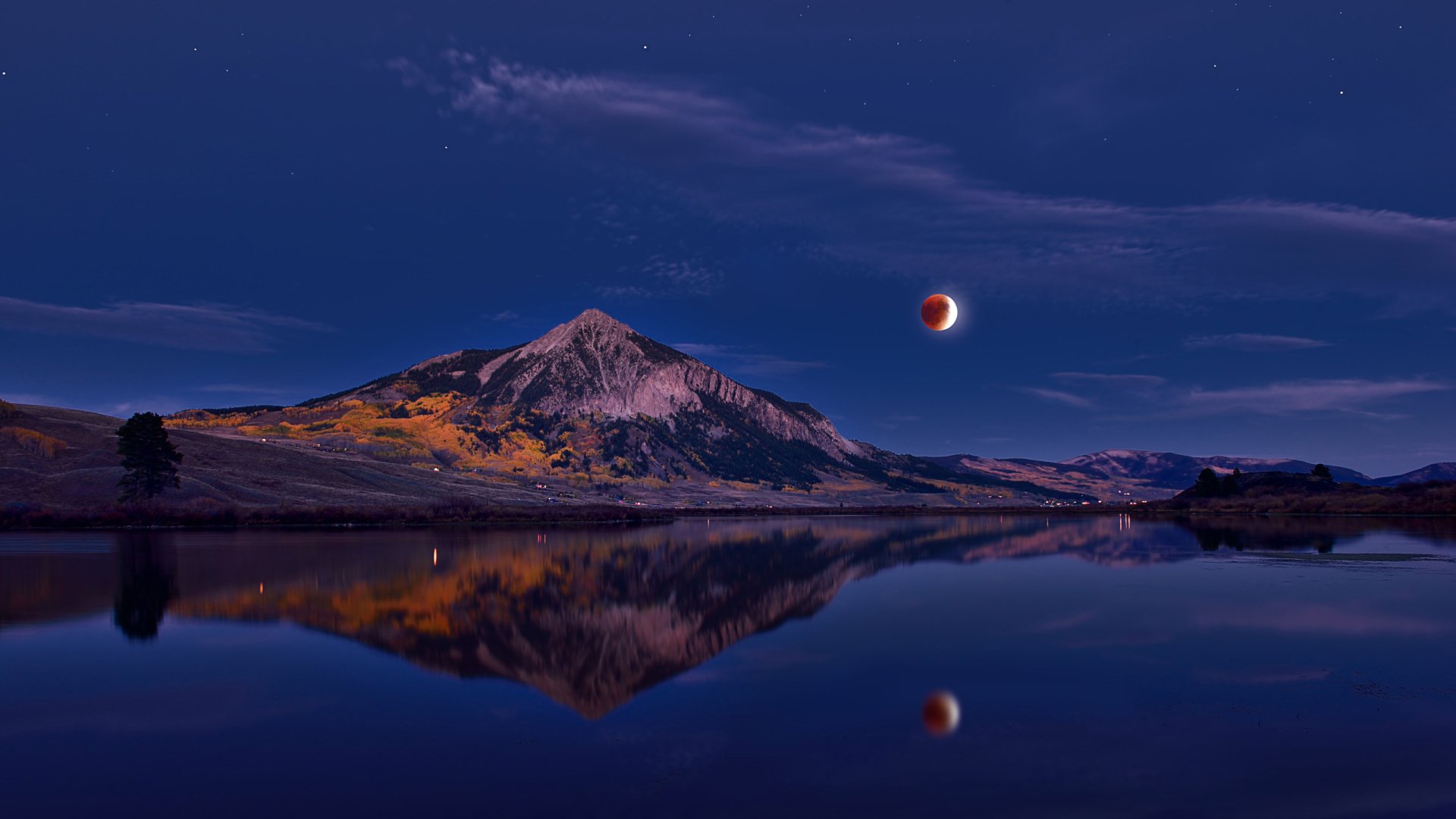 HD desktop wallpaper of a mountain landscape reflecting in calm water beneath a deep blue sky with a red lunar eclipse moon overhead.