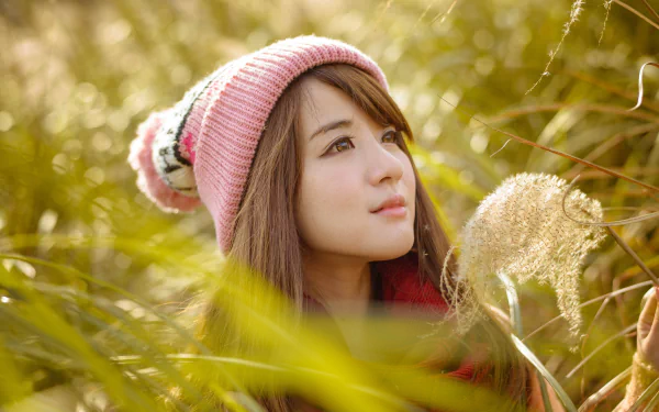 A 4K Ultra HD image of an Asian woman wearing a pink knit hat, surrounded by golden foliage in a serene outdoor setting.