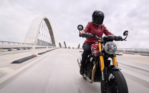A rider in a red jacket and helmet maneuvers a Triumph Speed 400 on a modern bridge, capturing the essence of freedom on two wheels in this HD wallpaper.