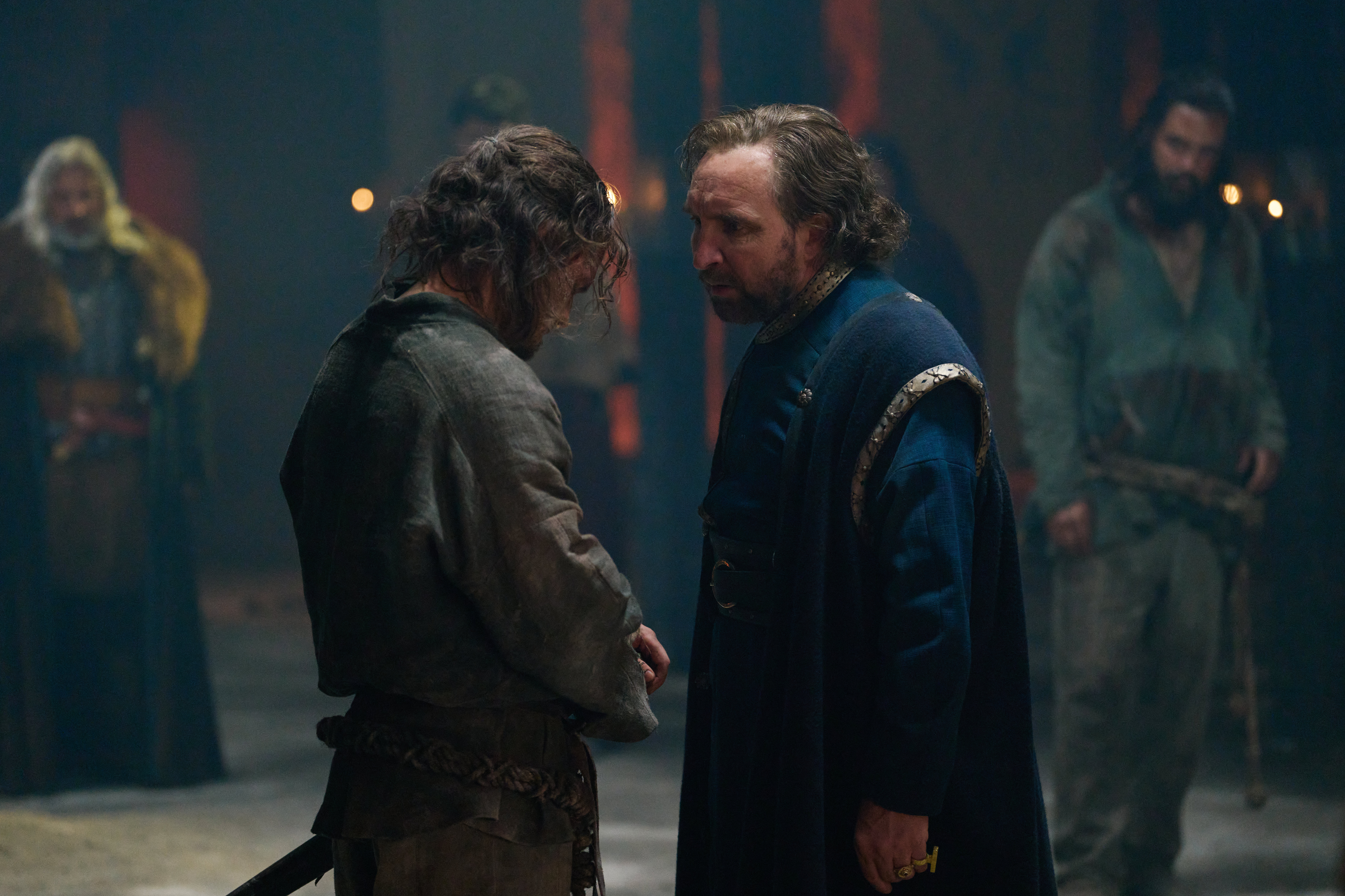 HD wallpaper featuring a scene with two characters from 'The Winter King', one, possibly Eddie Marsan, in a blue cloak engaging with another in a grim, medieval setting.