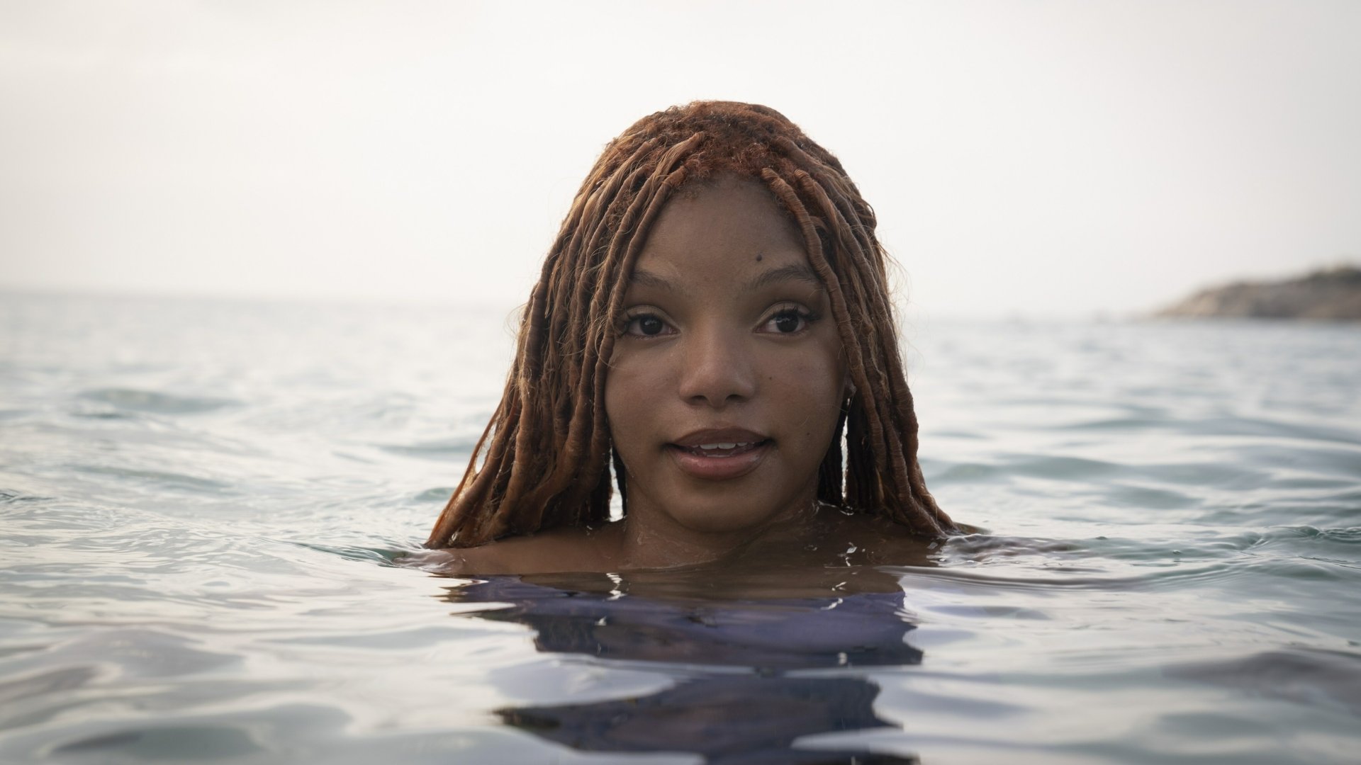 A serene HD background from the movie The Little Mermaid (2023), featuring a character with long hair, partially submerged in tranquil waters, exuding a sense of wonder and beauty.