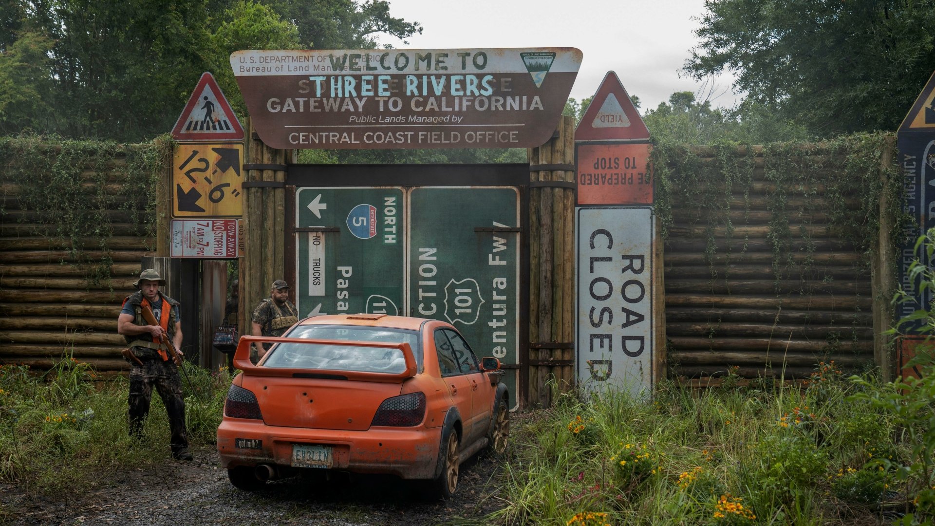 HD wallpaper of a scene from Twisted Metal showing an abandoned orange car in a rundown environment with signs indicating Three Rivers, set as a gloomy and atmospheric desktop background.