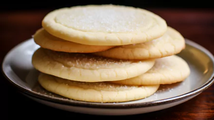 A close-up of a stack of sugar cookies on a plate, with a sprinkle of sugar on top, set against a warm wooden background, creating an inviting and appetizing desktop wallpaper.
