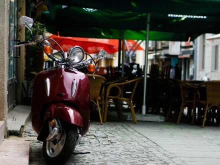 HD wallpaper of a red scooter parked on a cobblestone street beside a café with empty chairs and tables under green and red umbrellas.