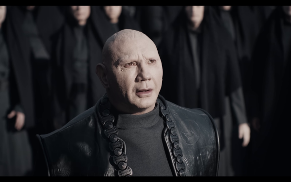 HD wallpaper featuring a character from Dune: Part Two, portrayed by an actor, standing with a solemn expression in front of a group of figures in dark attire.