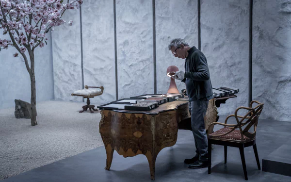 HD wallpaper featuring a scene from Interview with the Vampire with a character examining an object on an ornate table in a room with cherry blossoms and vintage furniture.