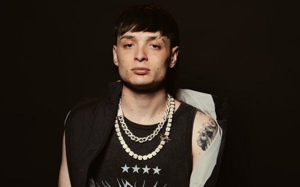 Portrait of a young man with a tattoo on his arm, wearing a black tank top with stars design and a gold necklace, embodying a Peso Pluma Mexican style, ideal for HD desktop wallpaper and backgrounds.