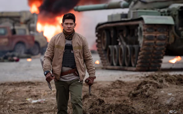 Iko Uwais strides confidently through a war-torn scene in Expend4bles, with flames and military vehicles in the background. This HD image serves as a striking desktop wallpaper.