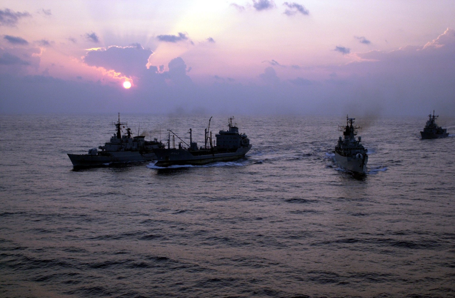 HD desktop wallpaper showing a military ship sailing on the ocean at sunset with a vibrant sky and calm waters.