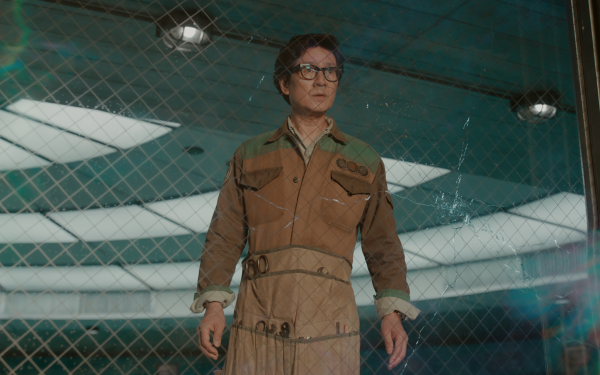 HD desktop wallpaper featuring a character inspired by Loki in a brown outfit standing confidently in front of a wire fence with a cracked glass pane.