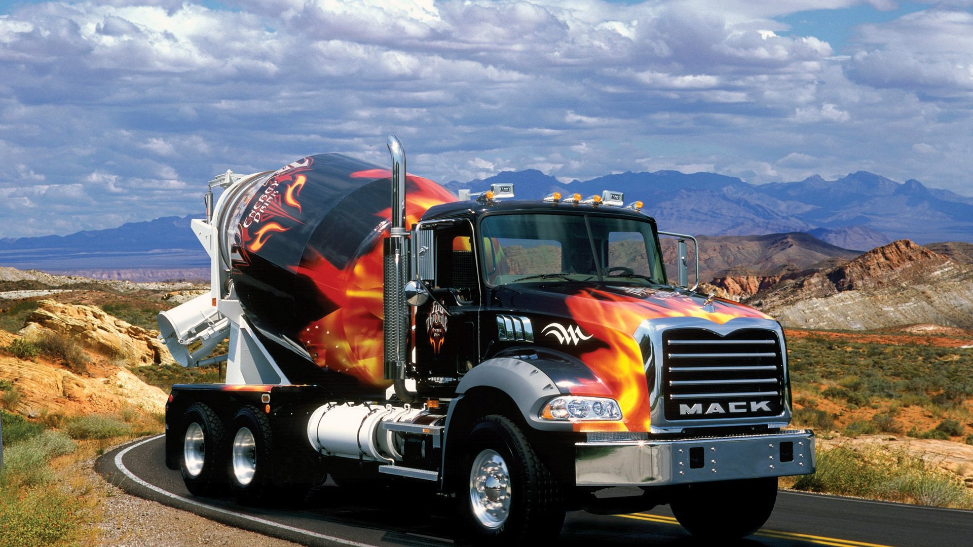 HD desktop wallpaper showcasing a Mack Trucks cement mixer with striking flame graphics, set against a scenic desert landscape under a partly cloudy sky.