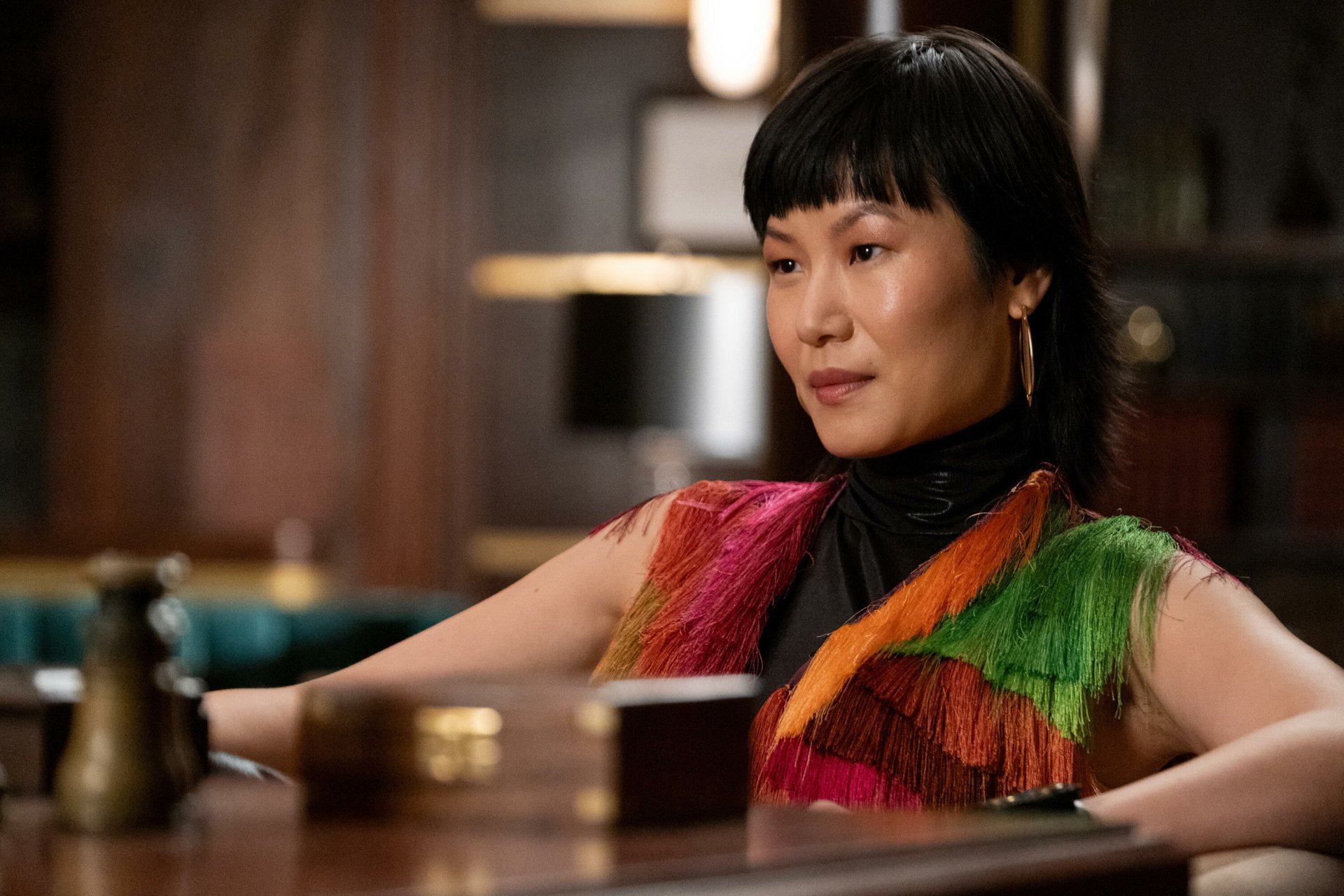 HD wallpaper featuring a scene from a TV show with a character contemplating Death and Other Details while sitting at a wooden table, donning a vibrant fringe outfit.