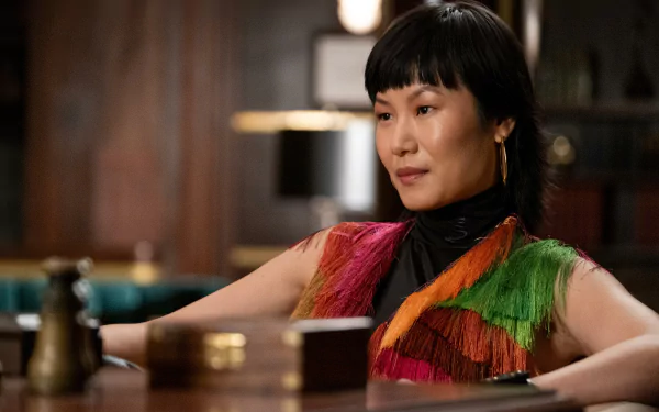 HD wallpaper featuring a scene from a TV show with a character contemplating Death and Other Details while sitting at a wooden table, donning a vibrant fringe outfit.