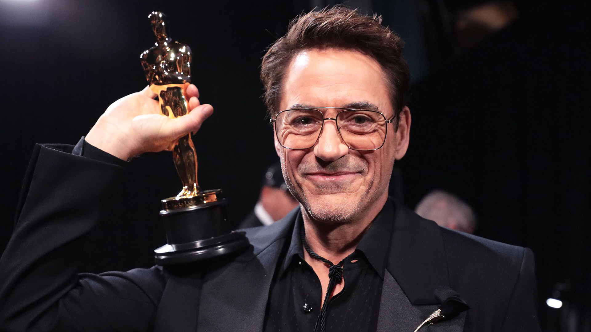 Smiling man in a black suit proudly holding an Academy Award (Oscar) trophy, suitable as a high-definition desktop wallpaper or background related to the Oscars.