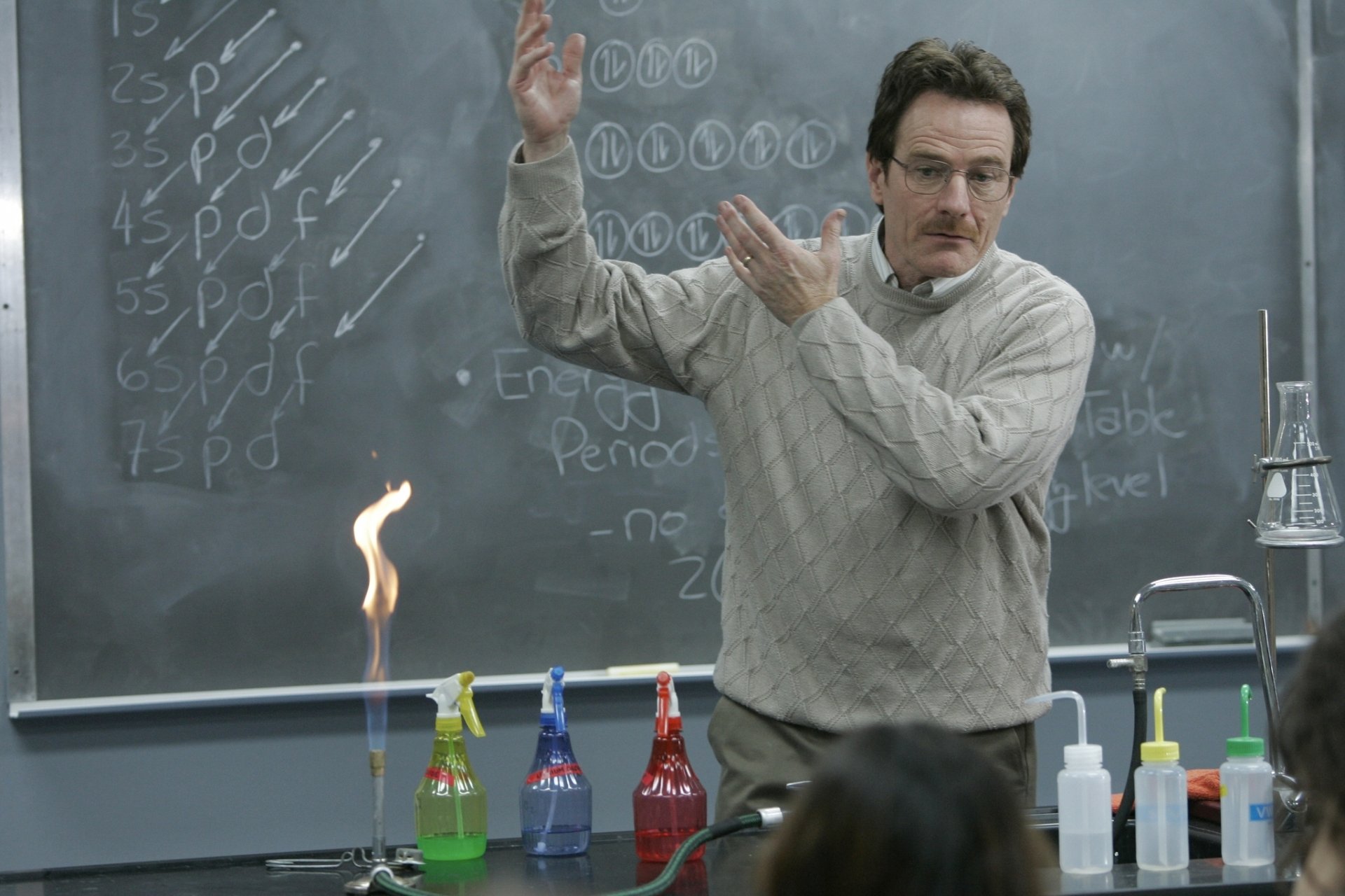 Walter White, portrayed by Bryan Cranston in the TV show Breaking Bad, passionately demonstrates a science experiment in a classroom setting. A flame rises from a colorful setup of bottles.