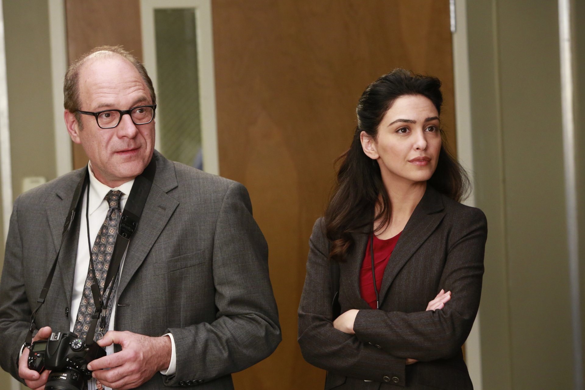 HD wallpaper featuring two characters from the TV show Grey's Anatomy standing in a hospital corridor, the man holding a camera and the woman with her arms crossed, both looking thoughtful.