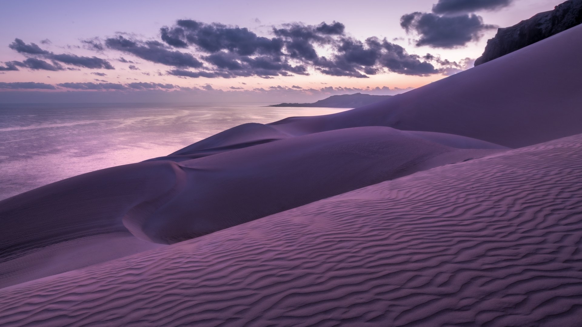 A serene landscape of rolling sand dunes under a twilight sky, featuring intricate patterns on the sand and soft, colorful clouds above. This HD image serves as a stunning desktop wallpaper.