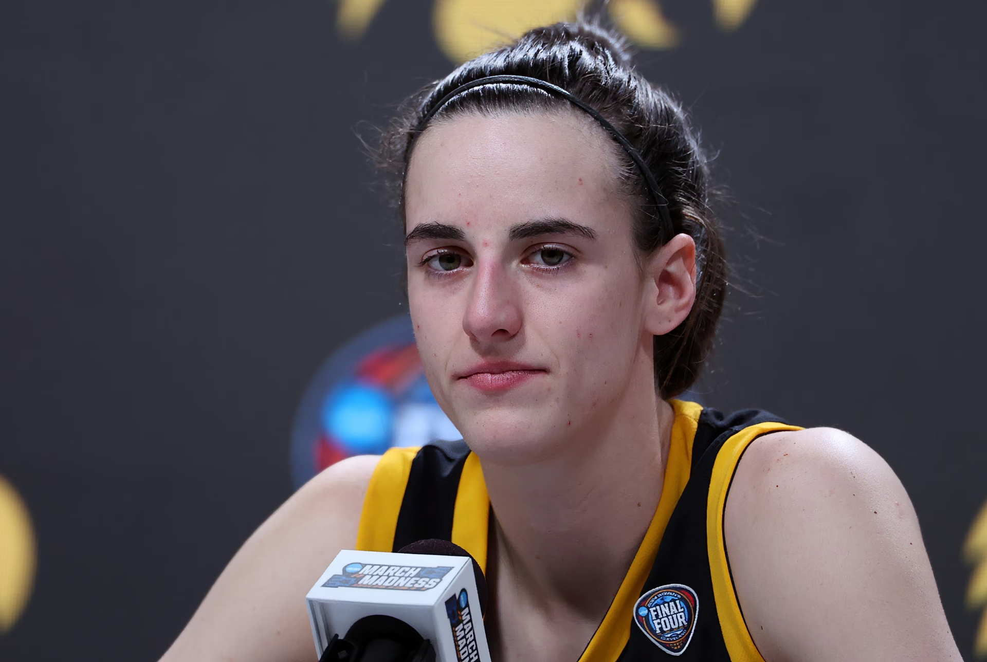 A basketball player in a black and gold jersey sits at a press conference with a focused expression, with a blurred background that includes the basketball logo. Suitable as an HD desktop wallpaper and background.