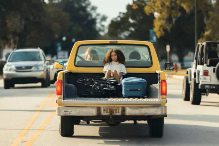 HD wallpaper from the movie Suncoast (2024) featuring a person sitting in the back of a pickup truck on a sunny road.