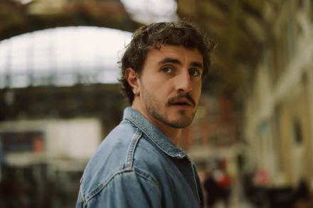 A cinematic still from the movie All of Us Strangers, featuring a man with curly hair and a denim jacket, set against a blurred, urban backdrop. An evocative HD desktop wallpaper.