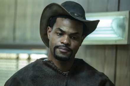 A character from The Walking Dead: The Ones Who Live wearing a cowboy hat and a brown coat, set against a backdrop of a rustic interior. HD wallpaper for fans of the show.