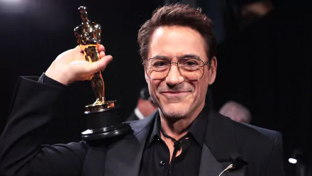 Smiling man in a black suit proudly holding an Academy Award (Oscar) trophy, suitable as a high-definition desktop wallpaper or background related to the Oscars.