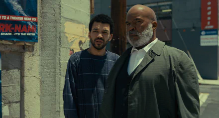 HD wallpaper featuring two actors from the TV show 'The American Society of Magical Negroes', with one dressed in a plaid shirt and the other in a grey suit, standing against an urban backdrop.