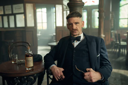 HD wallpaper featuring a scene from the TV show Peaky Blinders with a stylish man in a vintage suit sitting in an old-fashioned pub, with a whiskey bottle on the table, capturing the atmospheric setting of the series.