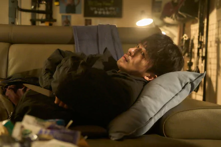 A man reclining on a couch with a book over his face, in a scene from the TV show Parasyte: The Grey, intended as an HD desktop wallpaper and background.