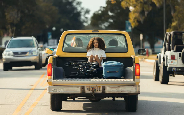 HD wallpaper from the movie Suncoast (2024) featuring a person sitting in the back of a pickup truck on a sunny road.