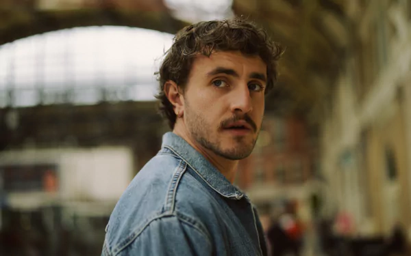 A cinematic still from the movie All of Us Strangers, featuring a man with curly hair and a denim jacket, set against a blurred, urban backdrop. An evocative HD desktop wallpaper.