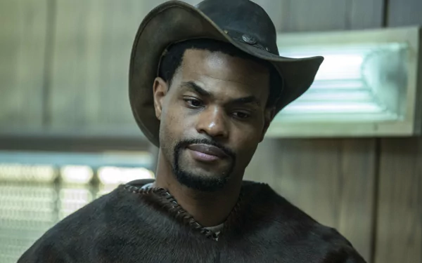A character from The Walking Dead: The Ones Who Live wearing a cowboy hat and a brown coat, set against a backdrop of a rustic interior. HD wallpaper for fans of the show.
