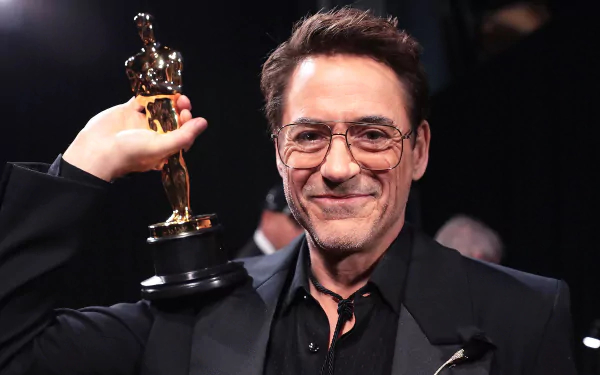 Smiling man in a black suit proudly holding an Academy Award (Oscar) trophy, suitable as a high-definition desktop wallpaper or background related to the Oscars.