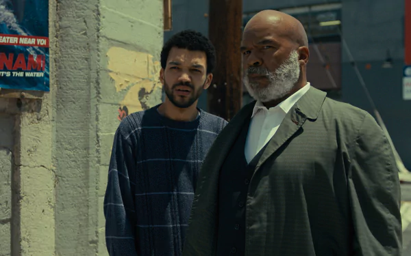HD wallpaper featuring two actors from the TV show 'The American Society of Magical Negroes', with one dressed in a plaid shirt and the other in a grey suit, standing against an urban backdrop.