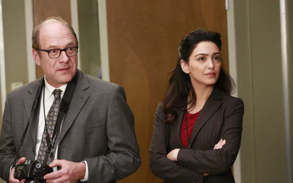 HD wallpaper featuring two characters from the TV show Grey's Anatomy standing in a hospital corridor, the man holding a camera and the woman with her arms crossed, both looking thoughtful.