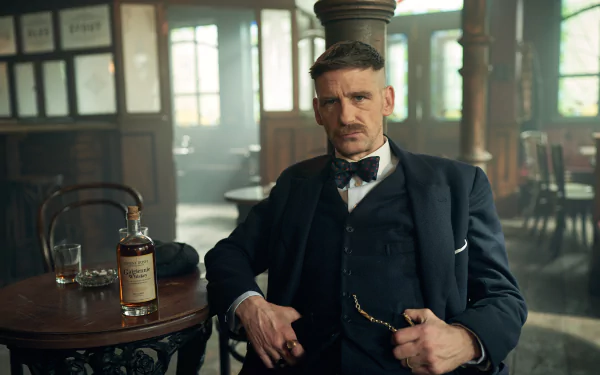 HD wallpaper featuring a scene from the TV show Peaky Blinders with a stylish man in a vintage suit sitting in an old-fashioned pub, with a whiskey bottle on the table, capturing the atmospheric setting of the series.