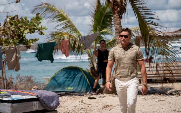 HD desktop wallpaper featuring a scene from the movie Road House (2024) with a focus on actor Billy Magnussen walking on a beach with palm trees and camping gear in the background.